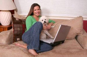 A woman sits with her small fluffy white dog in her lap on a couch with her laptop computer
